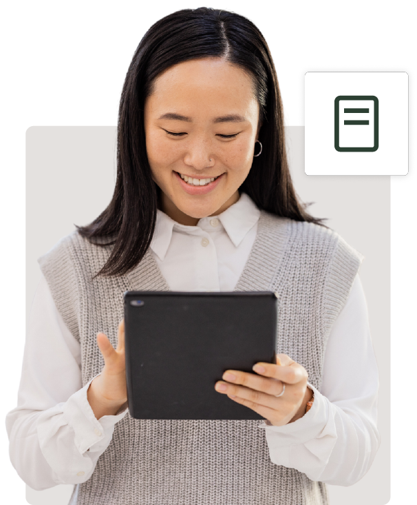 A young asian woman smiling, looking at a tablet she's holding with both hands, dressed in a white shirt and gray sweater vest, isolated on a white background.
