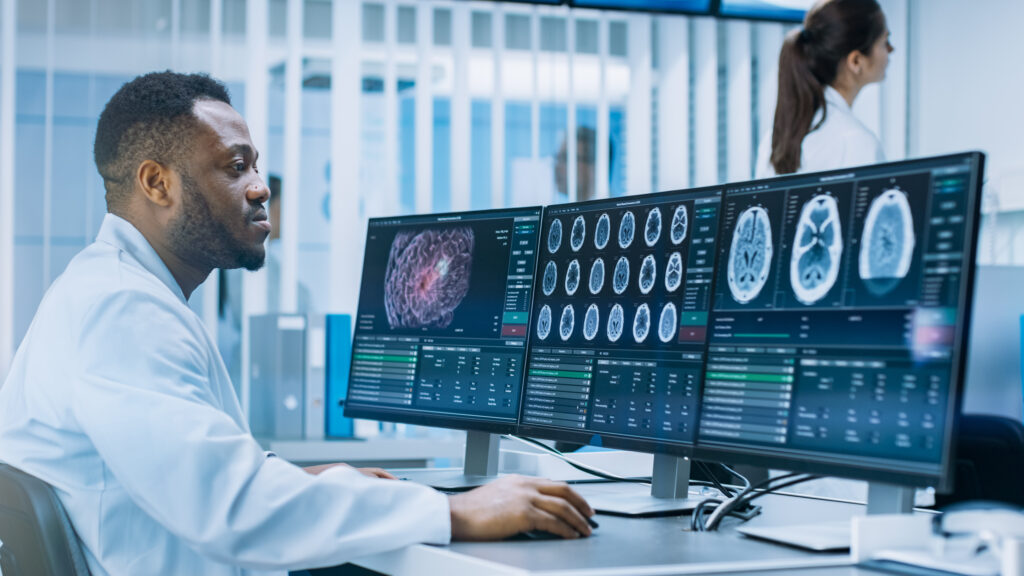 A male medical professional in a white lab coat is seated at a desk, analyzing brain scans displayed on two large computer monitors. In the background, another medical professional stands near a window with vertical blinds.