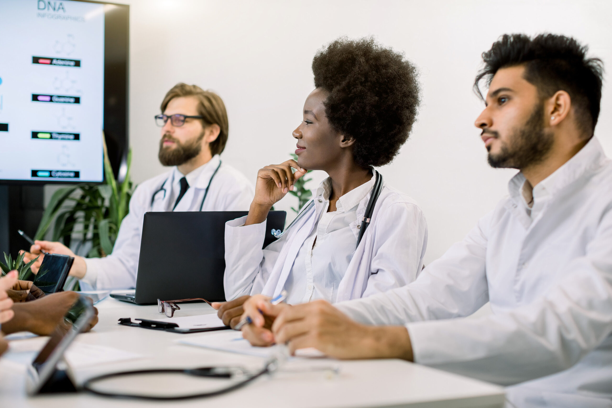 Three medical professionals in white coats are seated at a table during a meeting. They are focused and seem engaged in discussion. A whiteboard with charts and notes is partially visible in the background. Each person has a laptop or notebook in front of them.