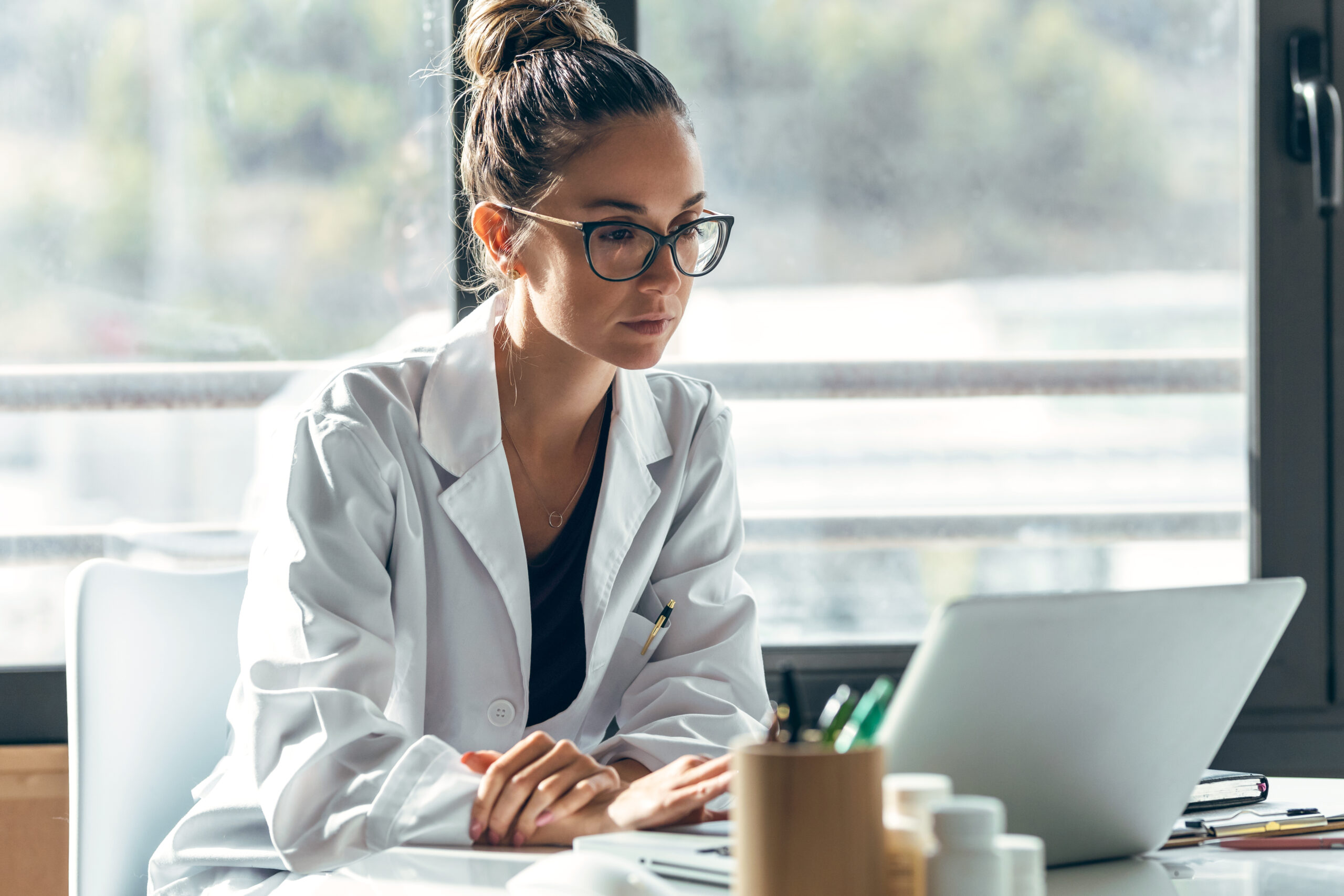 A woman in a white lab coat and glasses is sitting at a desk, working on a laptop. The background features large windows allowing natural light to fill the room. The desk has various office supplies and bottles on it.