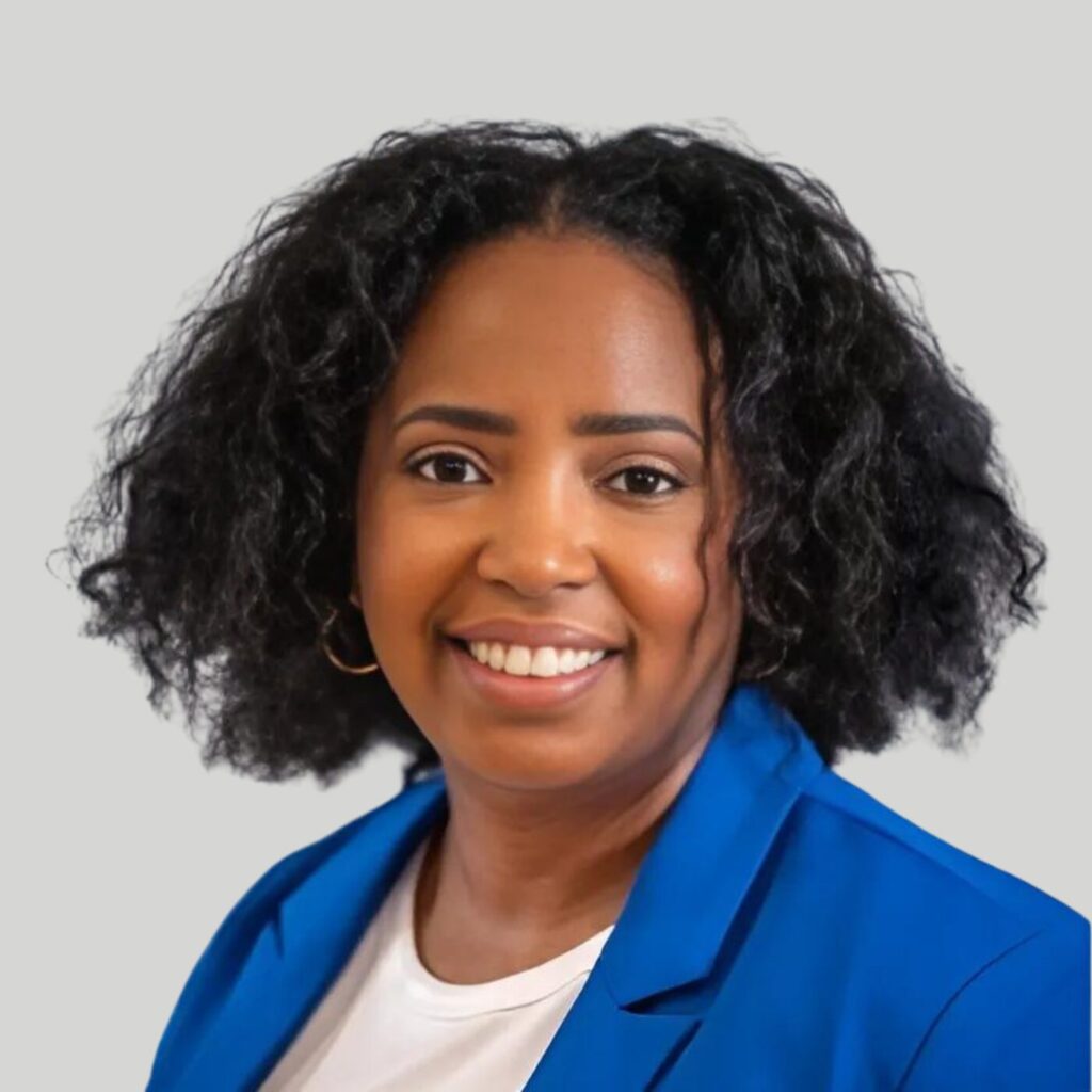 A woman with shoulder-length curly hair smiles at the camera. She is wearing a blue blazer over a white top, with gold hoop earrings. The background is plain and light grey.