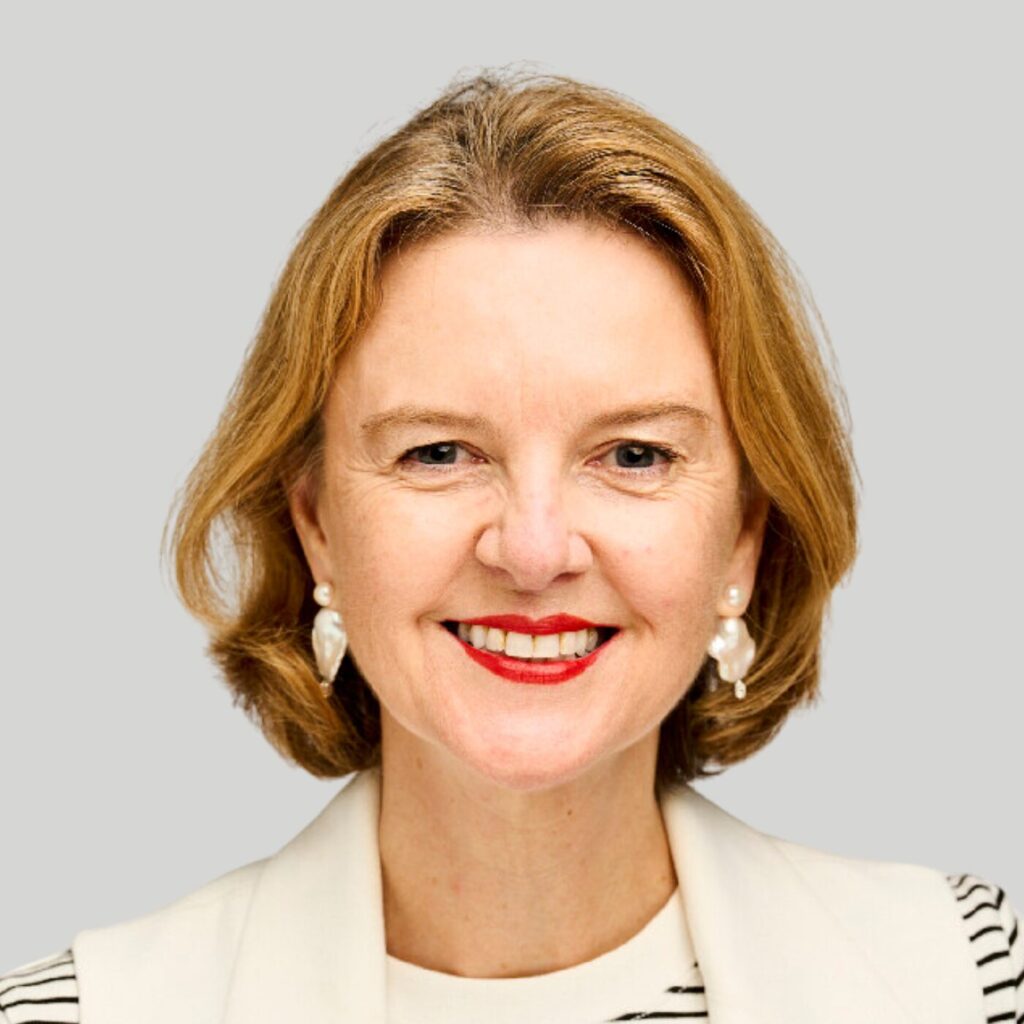 A woman with shoulder-length blonde hair is smiling at the camera. She is wearing pearl earrings, a white blazer, and a striped top. The background is gray.