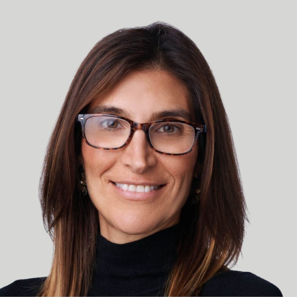 A woman with long brown hair and tortoiseshell glasses is smiling. She is wearing a black turtleneck against a light grey background.