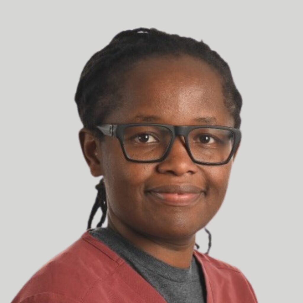 A person with short dreadlocks and large, rectangular glasses is smiling at the camera. They are wearing a maroon shirt and a gray inner shirt. The background is plain white.