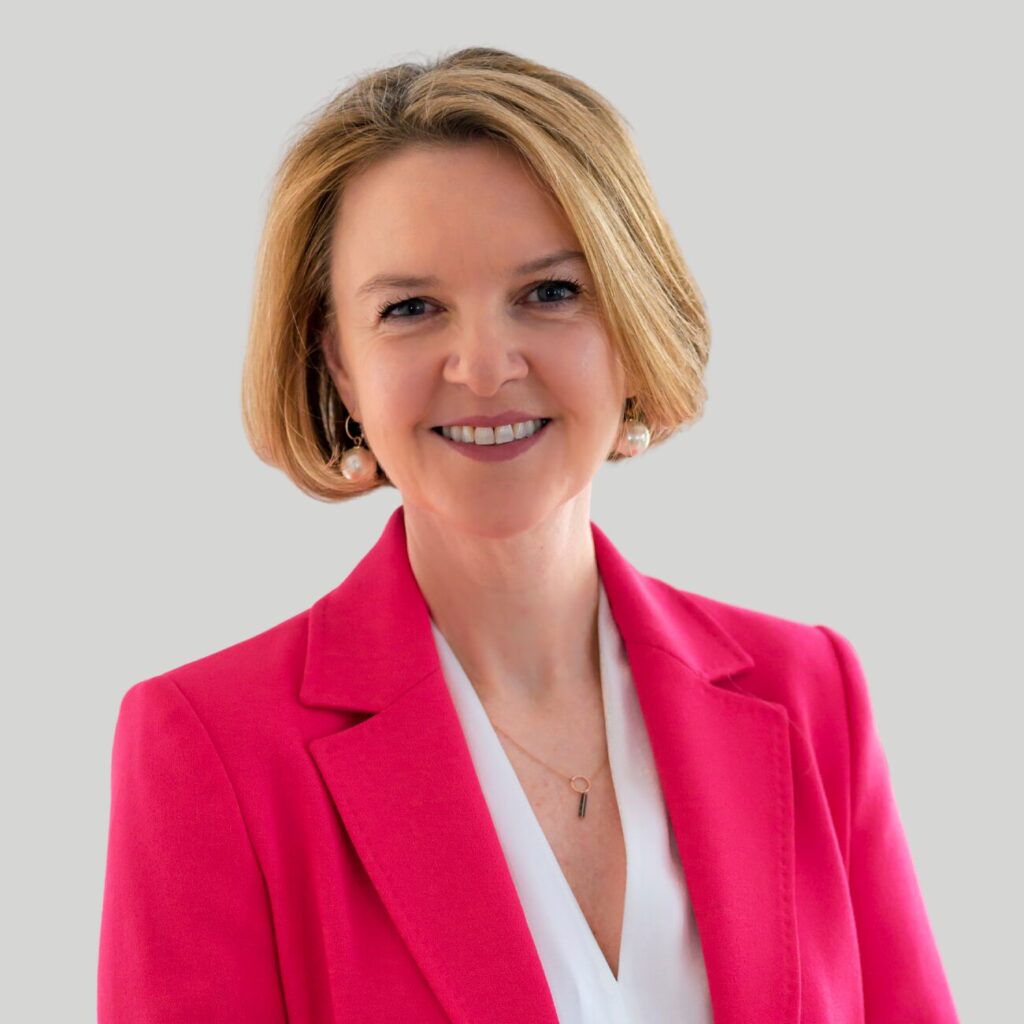 A person with short, blonde hair is smiling and wearing a bright pink blazer over a white blouse. The background is a plain, light gray color.